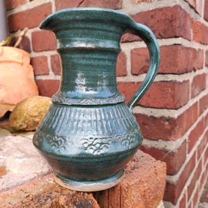 Könnte beinhalten: Eine türkisfarbene Keramikkrug mit gebogenem Griff und Ausguss. Der Krug hat einen bauchigen Boden und einen zylindrischen Hals mit dekorativen Rillen und floralen Mustern. Der Krug steht auf einer Ziegeloberfläche.