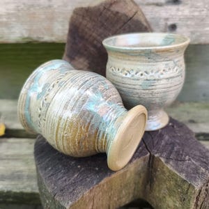 Puede incluir: Dos tazas de cerámica hechas a mano con un acabado texturizado y esmaltado. Las tazas son en tonos beige, azul y marrón. Una taza está tumbada de lado, mientras que la otra está de pie. Las tazas tienen una banda decorativa en el medio.