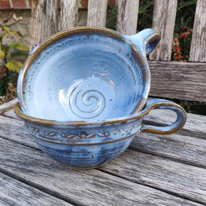 Può includere: Due ciotole in ceramica blu fatte a mano con manici, una impilata dentro l'altra. Le ciotole hanno una glassa blu chiaro con bordi marroni e un disegno a spirale al centro. Le ciotole sono su una superficie di legno.