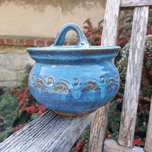 Könnte beinhalten: Ein runder, glasierter Keramiktopf mit glänzend blauer Oberfläche und passendem Deckel mit zentralem Bogengriff. Die Außenseite des Topfes ist mit einem sich wiederholenden braunen, wellenförmigen Muster verziert, das den Mittelteil umschließt.