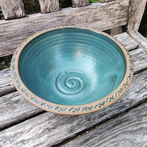 Op de afbeelding: Een ronde keramische kom met een glanzende blauwgroene binnenkant met een spiraalpatroon op de bodem. De buitenrand is van ongeglazuurde beige klei, versierd met een herhalend patroon van kleine, donkerblauwe streepjes en stippen rondom.