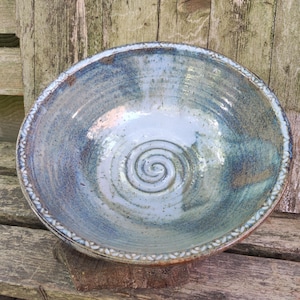 Puede incluir: Un cuenco de cerámica hecho a mano con un esmalte azul y verde. El cuenco presenta un diseño en espiral en el centro y un borde decorativo. El cuenco tiene aproximadamente 25 cm de diámetro.