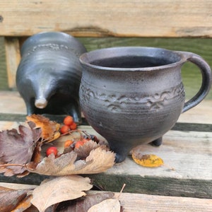 Op de afbeelding: Twee zwarte keramische mokken met een rustieke afwerking en drie poten. De mokken hebben een textuurontwerp en staan op een houten oppervlak met herfstbladeren en rode bessen.
