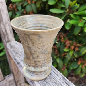 Può includere: Un calice in ceramica con bordo svasato e base a piedistallo. Il recipiente presenta una smaltatura beige e marrone screziata con sottili venature grigio-blu e creste orizzontali, rifinito con un motivo decorativo a festoni vicino alla base.