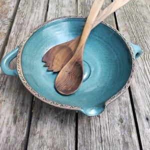 Puede incluir: Un cuenco de cerámica turquesa con asas y borde decorativo, que contiene dos cubiertos de ensalada de madera. El cuenco tiene una superficie texturizada y está sobre una mesa de madera envejecida. Una adición perfecta a cualquier cocina.