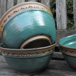Può includere: Ciotole in ceramica fatte a mano in turchese e marrone. Le ciotole hanno una finitura lucida e una fascia decorativa con un motivo ripetuto sul bordo. Le ciotole sono impilate, con una ciotola parzialmente visibile.