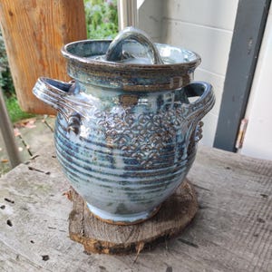Könnte beinhalten: Ein Keramikgefäß mit Deckel, das eine glänzende blau-braune Glasur mit horizontalen Rillen und einem geprägten Blumenmuster auf der Vorderseite aufweist. Das Gefäß hat zwei seitliche Griffe und einen kleinen Griff am Deckel sowie einen braunen Tonboden.