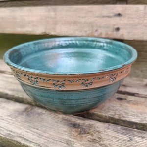 Op de afbeelding: Een handgemaakte keramische kom met een turquoise binnenkant en een bruine, versierde band rond de rand. De kom is rond en heeft een rustieke, getextureerde uitstraling.