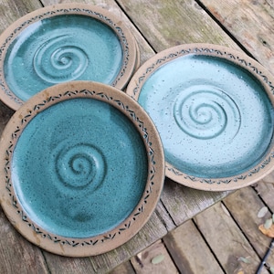 Könnte beinhalten: Drei handgefertigte Keramikplatten mit türkisfarbener Glasur und einem Spiraldesign in der Mitte. Die Teller haben einen dekorativen Rand und stehen auf einer Holzoberfläche. Eine einzigartige Ergänzung für jede Küche.