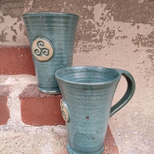 Può includere: Due tazze in ceramica color verde acqua con finitura strutturata. Ogni tazza presenta un emblema circolare in rilievo con un motivo a spirale. Una tazza ha un manico. Le tazze sono esposte su una superficie di mattoni.