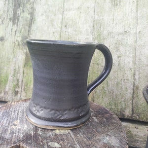 Könnte beinhalten: Eine schwarze Keramiktasse mit einer strukturierten Oberfläche und einem abgerundeten Griff. Die Tasse hat einen leicht ausgestellten Rand und einen breiten Boden.