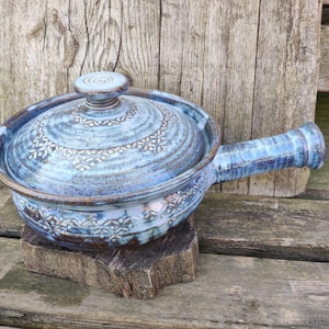 Op de afbeelding: Een blauwe keramische ovenschaal met deksel en handvat. De schaal heeft een decoratief bloemmotief en rust op een houten blok. De achtergrond is een houten oppervlak.