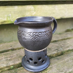 Może przedstawiać: Ciemnoszara ceramiczna śmietanka z uchwytem i wylewką, spoczywająca na pasującej podstawie z okrągłymi wycięciami. Śmietanka ma kwiatowy wzór wokół środka. Element znajduje się na drewnianej powierzchni.