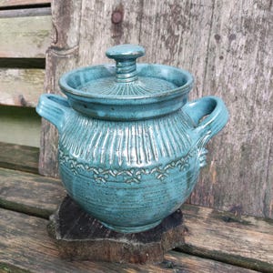 Puede incluir: Una olla de cerámica turquesa con tapa, asas y detalles decorativos tallados. La olla se coloca sobre una base de madera, mostrando una estética rústica y artesanal. El diseño de la olla incluye líneas talladas verticales y horizontales.