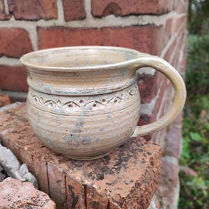 Könnte beinhalten: Eine handgefertigte Keramik-Tasse mit rundem Körper und großem Henkel. Die Tasse hat eine hellbraune Farbe mit einem dekorativen Band aus geätzten Mustern in der Mitte. Die Tasse steht auf einer Ziegeloberfläche.