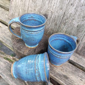 Può includere: Tre tazze in ceramica fatte a mano in tonalità di blu con accenti marroni. Ogni tazza ha un manico e un motivo di smalto unico. Le tazze sono esposte su una superficie di legno, mostrando il loro fascino rustico.