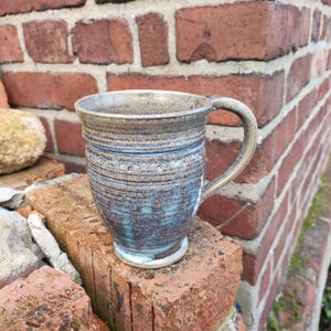 Könnte beinhalten: Eine handgefertigte Keramik-Tasse mit blau-brauner Glasur. Die Tasse hat einen abgerundeten Körper, einen stabilen Henkel und ein dekoratives Band am oberen Rand. Die Tasse steht auf einem roten Ziegelstein.