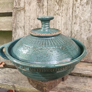 Op de afbeelding: Een turquoise keramische ovenschaal met een deksel. De schaal heeft een ronde vorm met handvatten en een decoratief bloemmotief op het deksel. Het deksel heeft een knopgreep. De schaal staat op een houten oppervlak.