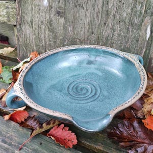 Puede incluir: Un cuenco de cerámica turquesa hecho a mano con un diseño en espiral en el centro. El cuenco tiene un borde beige claro con un patrón decorativo y dos asas pequeñas. El cuenco está sobre una superficie de madera con hojas de otoño.
