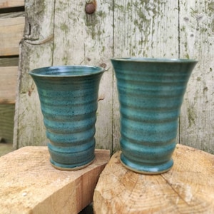 May include: Two teal blue ceramic cups with a textured, wavy design. The cups are sitting on a wooden surface.