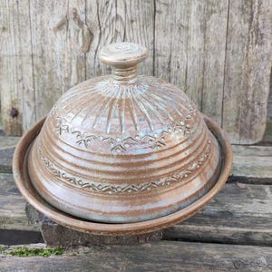 Könnte beinhalten: Eine handgefertigte Butterdose aus Keramik mit Deckel. Die Schale hat eine warme braune Farbe mit einem dekorativen Muster um den Boden und den Deckel. Der Deckel hat einen kleinen, runden Griff oben. Die Schale steht auf einer Holzoberfläche.