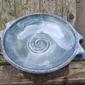 Puede incluir: Un cuenco de cerámica azul hecho a mano con un diseño en espiral en el centro. El cuenco tiene dos asas y un borde decorativo. El cuenco tiene aproximadamente 30 cm de diámetro.