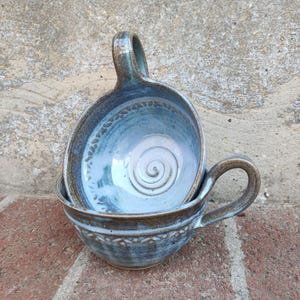 Può includere: Due ciotole in ceramica fatte a mano con manici, una impilata nell'altra. Le ciotole sono smaltate in tonalità di blu e marrone, con una spirale decorativa al centro della ciotola superiore. I bordi presentano un motivo strutturato.