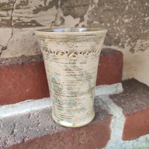Puede incluir: Un vaso de cerámica hecho a mano con un acabado beige claro texturizado y una banda decorativa de diseños recortados cerca de la parte superior. La taza tiene una forma ligeramente acampanada y mide unos 15 cm de alto.