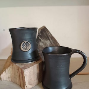 Op de afbeelding: Twee zwarte keramische mokken met een rustieke uitstraling. Eén mok heeft een Keltisch knoopontwerp. De mokken zijn cilindrisch met een uitlopende basis en een handvat. De mokken worden op een houten oppervlak gepresenteerd.
