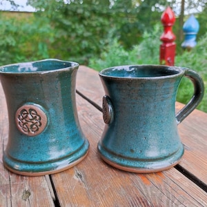 Könnte beinhalten: Zwei tealfarbene Keramikbecher mit einem keltischen Knotendesign. Die Becher haben eine braune Glasur und eine leicht strukturierte Oberfläche.
