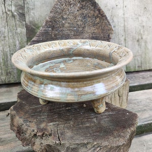 Op de afbeelding: Een ronde, driepotige keramische kom met een brede, platte rand voorzien van gegraveerde runensymbolen. De kom heeft een glanzend, gevlekt glazuur in tinten beige, bruin en saliegroen, met zichtbare concentrische ringen op de bodem van de kom.