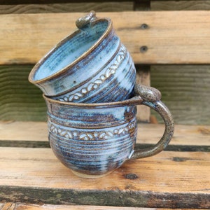 Könnte beinhalten: Zwei handgefertigte Keramikbecher mit blauer und brauner Glasur. Die Becher haben ein weißes Design um den oberen und unteren Rand. Die Becher sind übereinander gestapelt.