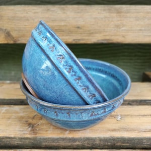 Könnte beinhalten: Zwei türkisfarbene Keramikschalen, übereinander gestapelt, die das detaillierte Randdesign zeigen. Die Schalen sind glasiert und stehen auf einer Holzoberfläche. Die Farbe und das Design der Schalen unterstreichen die handwerkliche Kunstfertigkeit.