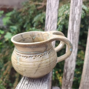 Può includere: Una tazza in ceramica beige di forma rotonda con un manico ampio e curvo e una superficie testurizzata con scanalature. La tazza presenta una fascia decorativa con piccole rientranze circolari sulla parte superiore, con sottili riflessi di smalto verde e marrone.