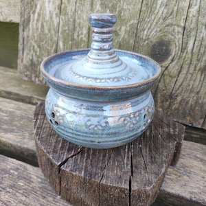 Pode incluir: Um pote de cerâmica artesanal com tampa, apresentando um esmalte azul claro com detalhes em azul escuro. O pote tem uma base redonda com orifícios decorativos e está sobre uma superfície de madeira. A tampa tem uma alça cilíndrica alta.