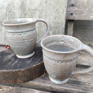 May include: Two ceramic mugs with a light brown and gray glaze. The mugs have a textured, rustic look and feature a decorative pattern around the rim.