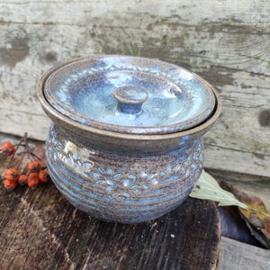 Könnte beinhalten: Eine handgefertigte Keramik-Butterdose mit Deckel. Die Dose hat eine Mischung aus Blau- und Brauntönen, eine strukturierte Oberfläche und ein dekoratives Blumenmuster am Boden. Der Deckel hat einen kleinen Knopf.