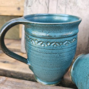 May include: A teal ceramic mug with a curved handle and a decorative band of embossed floral designs. The mug has a textured surface and is approximately 10 cm tall. The mug is part of a set, with other mugs visible in the background.