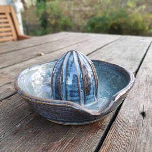 May include: A handmade ceramic citrus juicer with a blue and brown glaze. The juicer has a ridged reamer and a bowl with a pouring spout. The pottery is sitting on a wooden table.