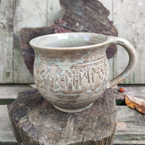 Könnte beinhalten: Eine Keramiktasse mit brauner und weißer Glasur. Die Tasse hat einen abgerundeten Körper und einen Henkel. Die Tasse hat Runen, die in die Seite geschnitzt sind.