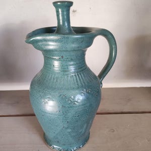 Może przedstawiać: Turkusowy ceramiczny dzbanek z uchwytem i wylewką. Dzbanek ma unikalny design z podniesionym środkiem i dekoracyjnym wzorem kwiatowym w pobliżu podstawy. Dzbanek ma około 25 cm wysokości.