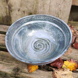 Puede incluir: Un cuenco de cerámica hecho a mano con interior azul claro y borde azul más oscuro. El cuenco presenta un diseño en espiral en el centro y está rodeado de hojas de otoño sobre una superficie de madera. El cuenco tiene unos 25 cm de diámetro.