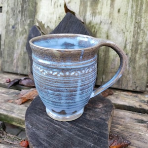Könnte beinhalten: Eine blaue und braune Keramiktasse mit texturierter Glasur und einem dekorativen Band am oberen Rand. Die Tasse hat einen Henkel und steht auf einer Holzoberfläche.