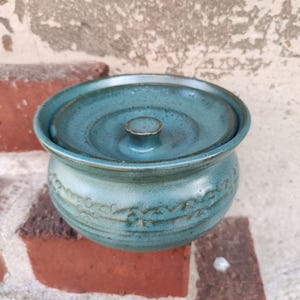 Könnte beinhalten: Eine türkisfarbene Butterdose aus Keramik mit Deckel. Die Dose hat eine runde Form mit einem dekorativen Blumenmuster am Boden. Der Deckel hat einen kleinen Knopf in der Mitte. Die Dose steht auf einer Ziegeloberfläche.