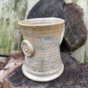 Puede incluir: Una taza de cerámica hecha a mano con un esmalte marrón claro y un diseño azul y blanco. La taza tiene un cuerpo redondeado y un asa. La taza está sentada sobre una superficie de madera.