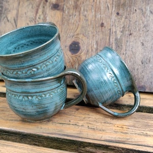 Può includere: Tre tazze in ceramica turchese con manici, una inclinata. Le tazze hanno una finitura strutturata e una fascia decorativa con motivi floreali in rilievo. Sono appoggiate su una superficie di legno.