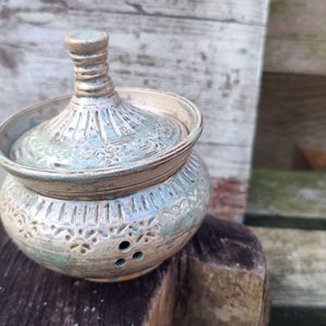 Op de afbeelding: Een handgemaakte keramische pot met een deksel. De pot is lichtblauwgroen van kleur met ingewikkelde bloempatronen en kleine gaatjes. Het deksel heeft een geribbeld ontwerp en een kleine knop. De pot staat op een houten oppervlak.