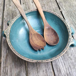Puede incluir: Un cuenco de cerámica turquesa con dos cucharas de ensalada de madera. El cuenco tiene un borde y asas decorativas. Las cucharas son de madera de color claro. El cuenco y las cucharas están sobre una superficie de madera envejecida.
