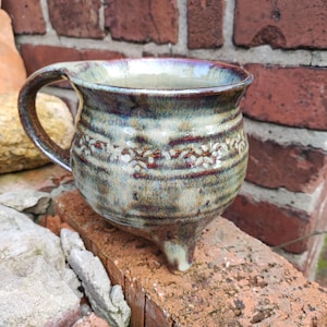 Könnte beinhalten: Eine handgefertigte Keramiktasse mit einem einzigartigen Design. Die Tasse hat einen abgerundeten Körper, einen Henkel und drei kleine Füße. Die Außenseite ist mit einer marmorierten Glasur in Blau-, Braun- und Cremetönen versehen, mit einem dekorativen Band in der Mitte.