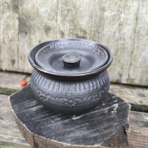 May include: A dark grey ceramic butter dish with a lid. The dish has a round shape with a decorative floral pattern around the base. The lid has a small knob and a matching floral design. The dish is sitting on a wooden surface.